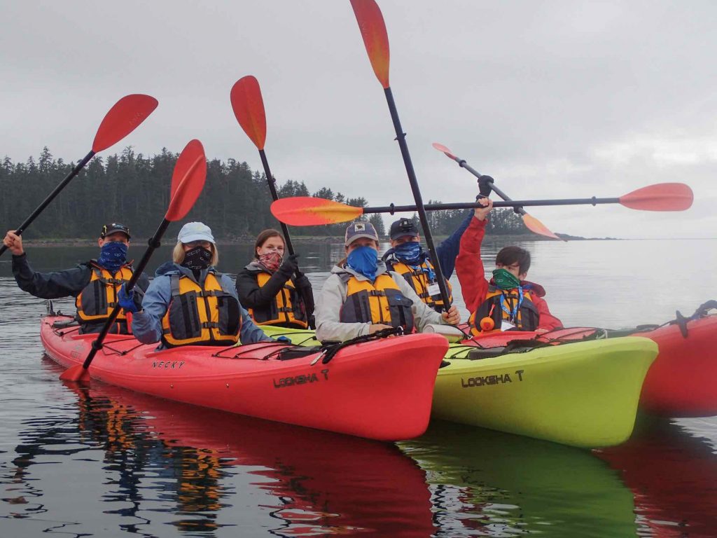 在阿拉斯加，UnCruise的客人们在皮划艇上戴着面具，把桨举在空中。©UnCruise