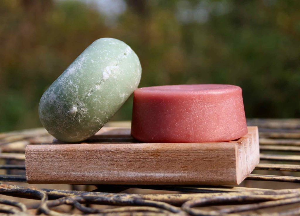 绿色和粉色的洗发水和护发素条放在户外的肥皂盘上，以绿色自然为背景。固体洗漱用品是可持续旅行的一个简单组成部分。开云体育总入球