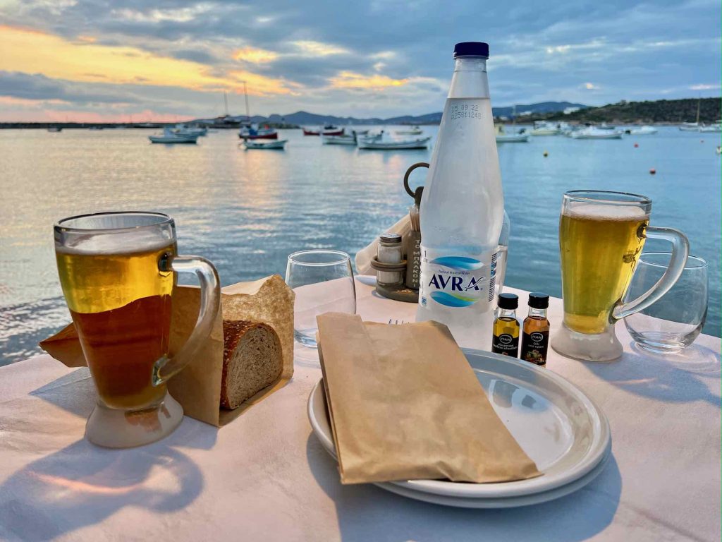 在希腊帕罗斯岛的一家海边餐厅吃晚餐，背景是地中海上的帆船，有一个塑料水瓶、面包、纸袋里的银器和一次性塑料单份橄榄油。©开云体育登录入口手机版官网网址KettiWilhelm2021