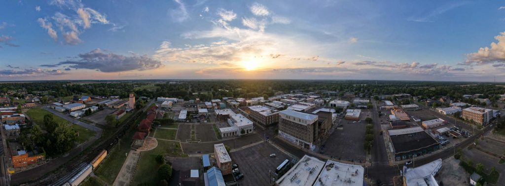 密西西比州克拉克斯代尔市中心日落时分的航拍。照片左边是一列货运火车，一到五层楼高的砖砌建筑覆盖了小镇的其余部分，而太阳在中心的地平线上盘旋。©ChristineLozada2022